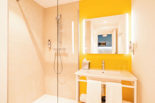 A modern bathroom with a glass shower enclosure, a white sink, and a yellow accent wall. - Appartements Soléo Prado (Marseille)