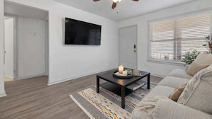 Ein Wohnzimmer mit Deckenventilator, einem flachbildfernseher an der Wand, einem Couchtisch mit Kerze und einem beige-farbenen Sofa.