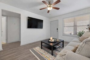 A living room with a ceiling fan, a flat-screen TV mounted on the wall, a coffee table with a candle, and a beige sofa. - Walk to AT&T Stadium • 2-Bedroom • Free Parking (Arlington)