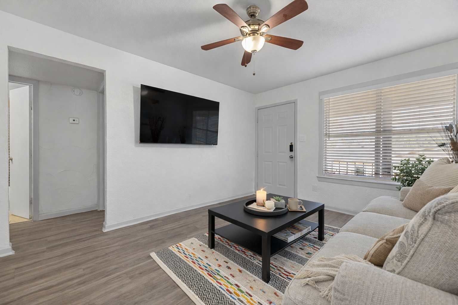 A living room with a ceiling fan, a flat-screen TV, a coffee table with a candle, a sofa, and a patterned rug.