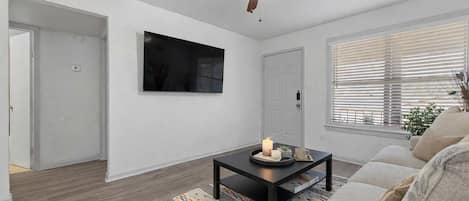 Ein Wohnzimmer mit Deckenventilator, einem Flachbildfernseher, einem Couchtisch mit Kerze, einer Couch und einem gemusterten Teppich.