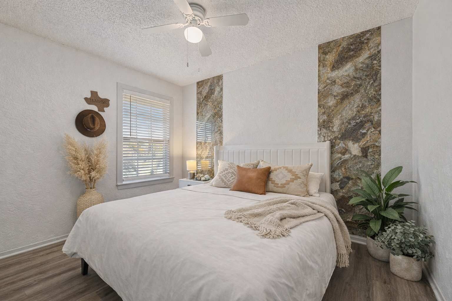 A bedroom with a bed, a ceiling fan, a window with blinds, a wall-mounted hat, and a vase with dried plants.