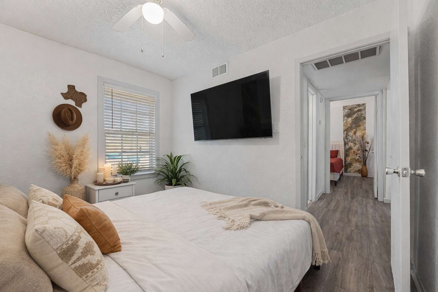 A bedroom with a bed, bedside table, lamp, wall-mounted TV, and a door leading to another room.