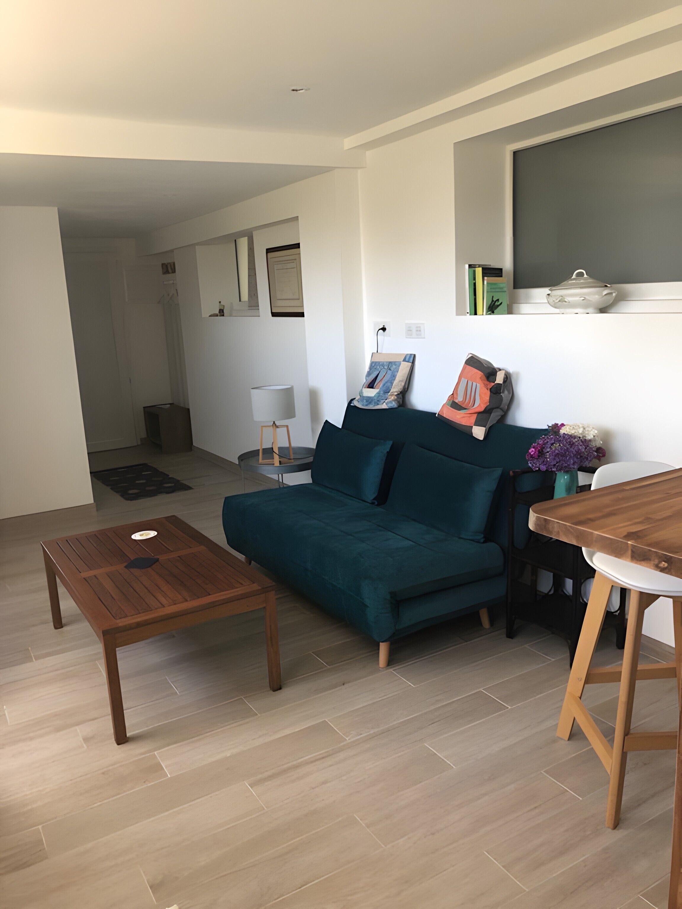 A living room with a teal sofa, a wooden coffee table, a dining table, and a vase with flowers.