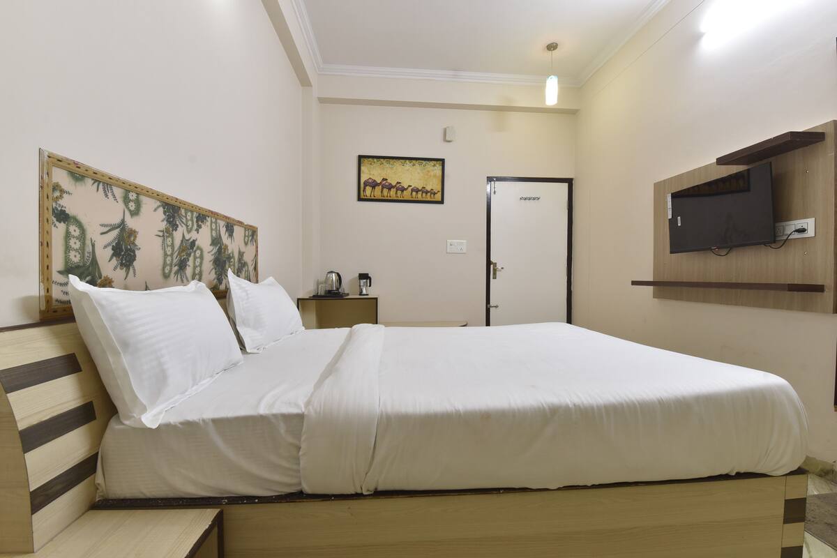 A neatly made bed with white linens, a wooden headboard, a small bedside table with a lamp, a wall-mounted TV, and a door with a glass panel.