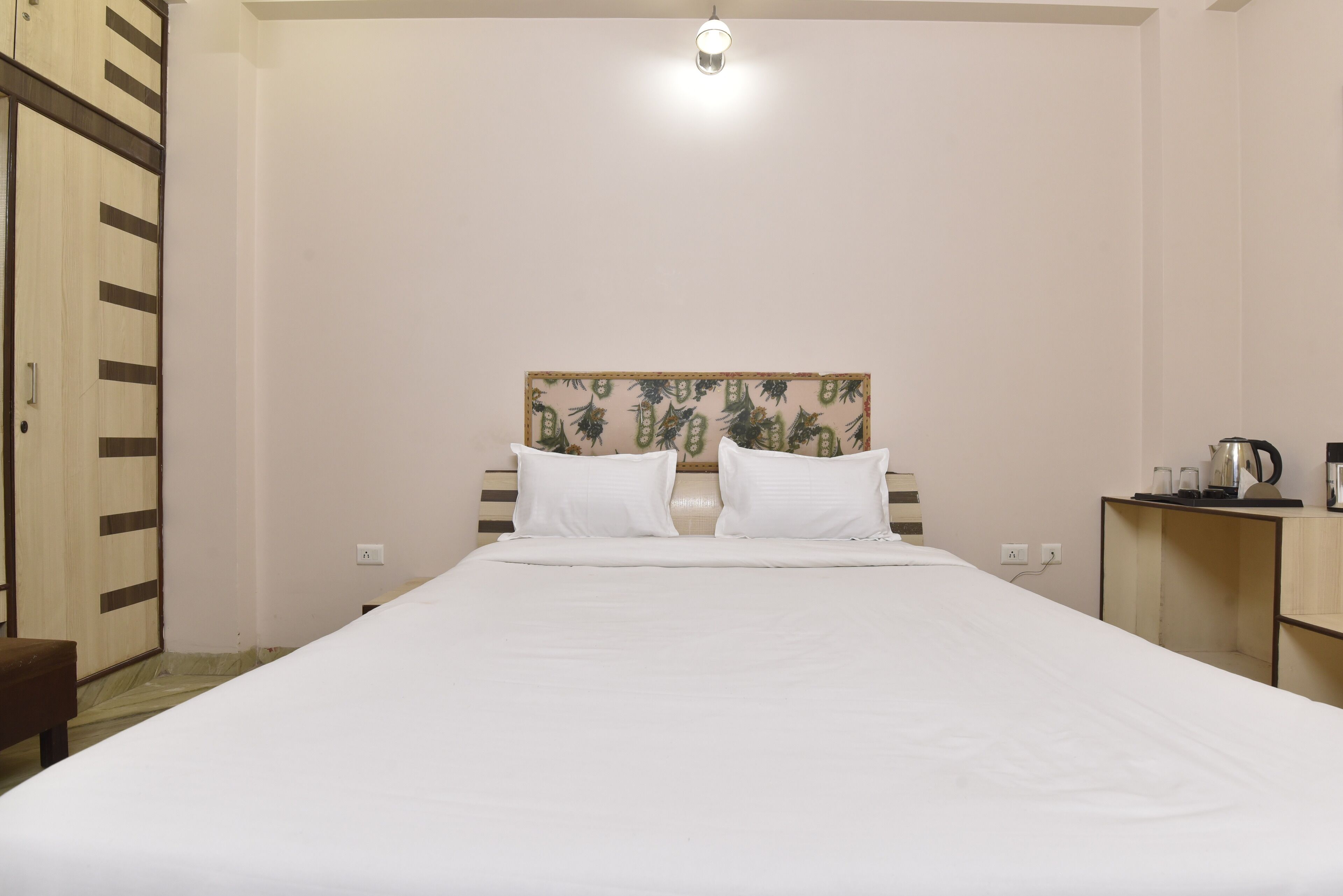 A hotel room with a large bed, a wooden headboard, a bedside table with a kettle, and a small cabinet.