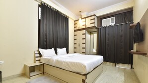 A bedroom with a bed, a small bedside table, a grey rug, a wooden wardrobe, a TV mounted on the wall, and a window with curtains. - Hotel The Grand Kailash (Jaipur)