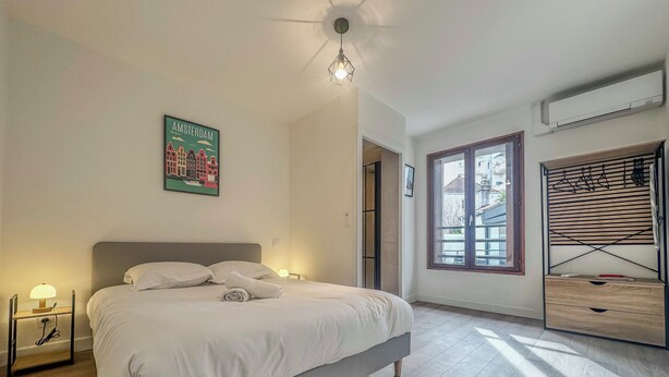 A bedroom with a bed, a nightstand, a wall-mounted air conditioner, and a window with a view of buildings. - #IM | The Victoria 10 💟 (Grenoble)