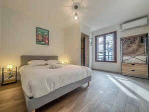 Un dormitorio con cama, mesita de noche, aire acondicionado de pared y ventana con vista a edificios.