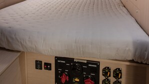 Una cama con un panel de control debajo, una ventana con vista al mar y un estante con objetos.