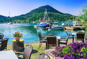 Ein Restaurant am Wasserufer mit Außenbestuhlung, in der Nähe liegen Boote vor Anker und im Hintergrund erhebt sich ein Berg.
