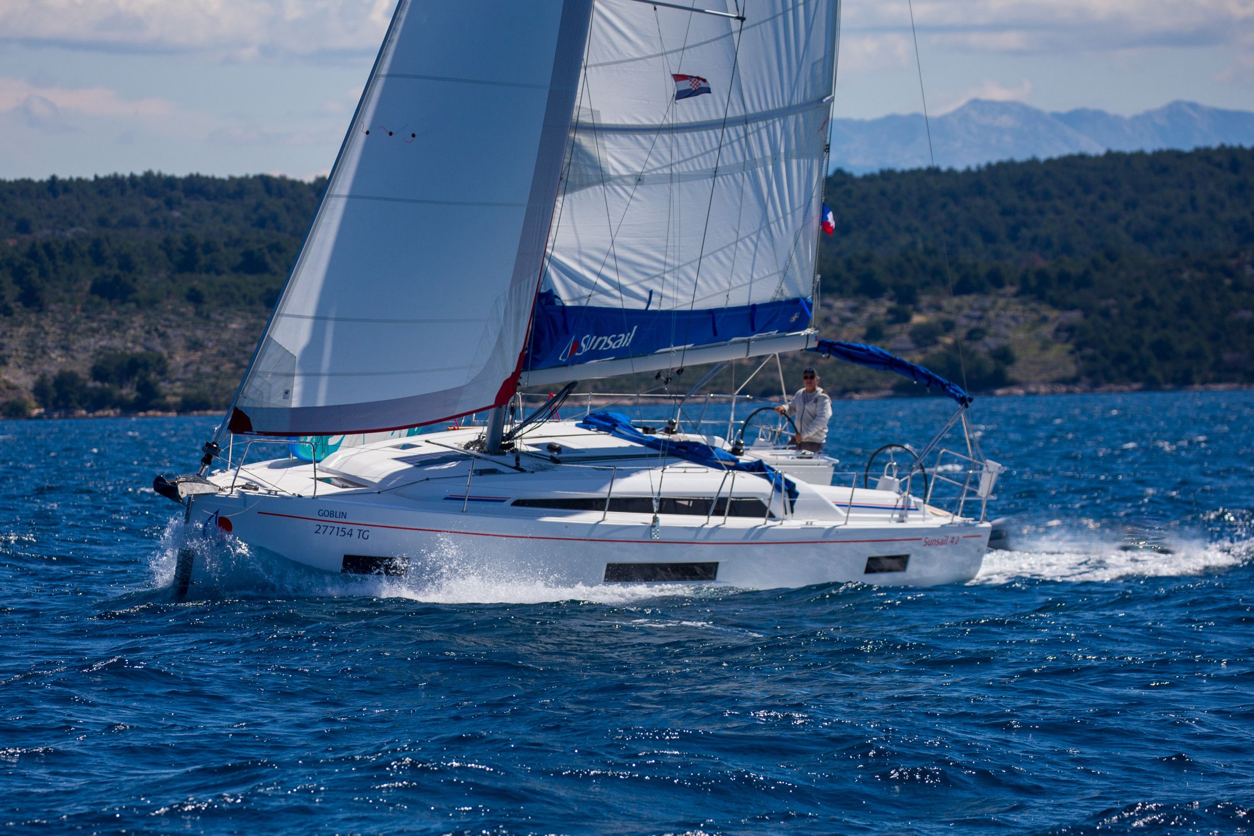 A sailboat with a cabin and a person standing at the helm, sailing on a body of water with a mountainous landscape in the background.