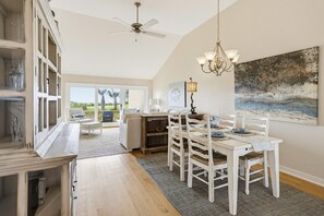 Ein Essbereich mit einem weißen Tisch, Stühlen und einer Chandelier. Es gibt einen Deckenventilator, einen Blick ins Freie und ein Bild an der Wand.