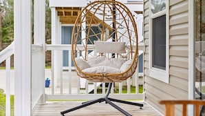Un fauteuil suspendu en osier, doté de coussins, est fixé à une rambarde sur une terrasse en bois.