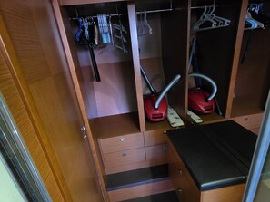 Un placard en bois contenant des vêtements suspendus, deux aspirateurs et un banc noir.