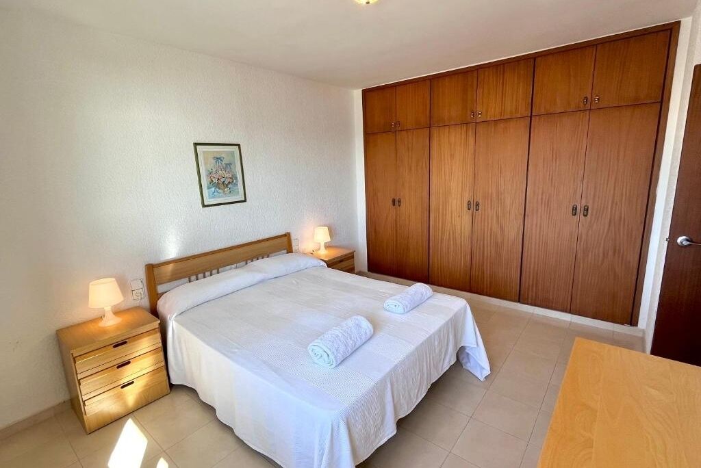 A bedroom with a wooden wardrobe, a bed with white bedding, and a nightstand with a lamp.