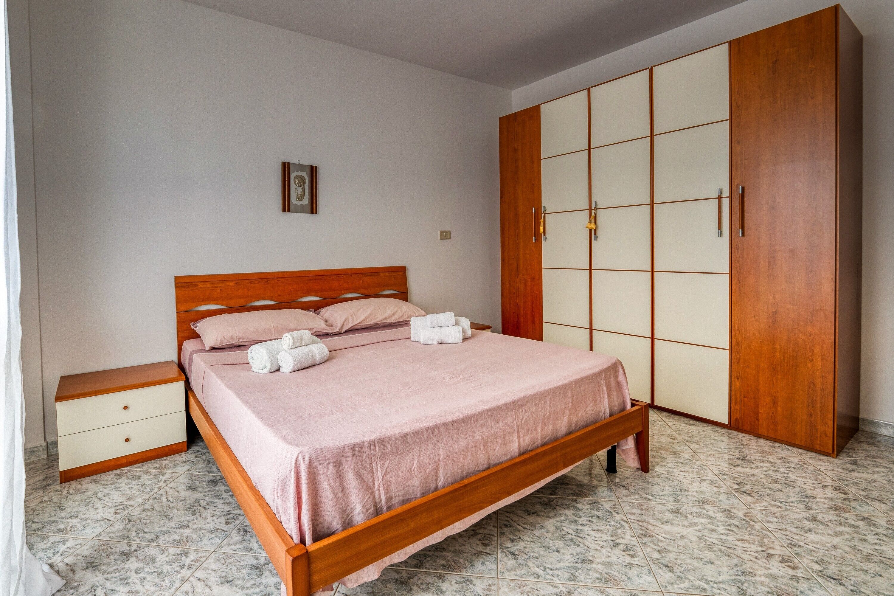 A bedroom with a wooden bed, a nightstand, a wardrobe, and a tiled floor.