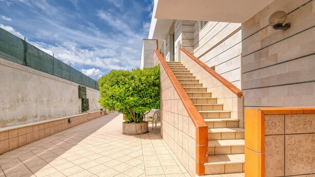 Un couloir au sol carrelé, un escalier avec des rampes orange et une plante en pot.