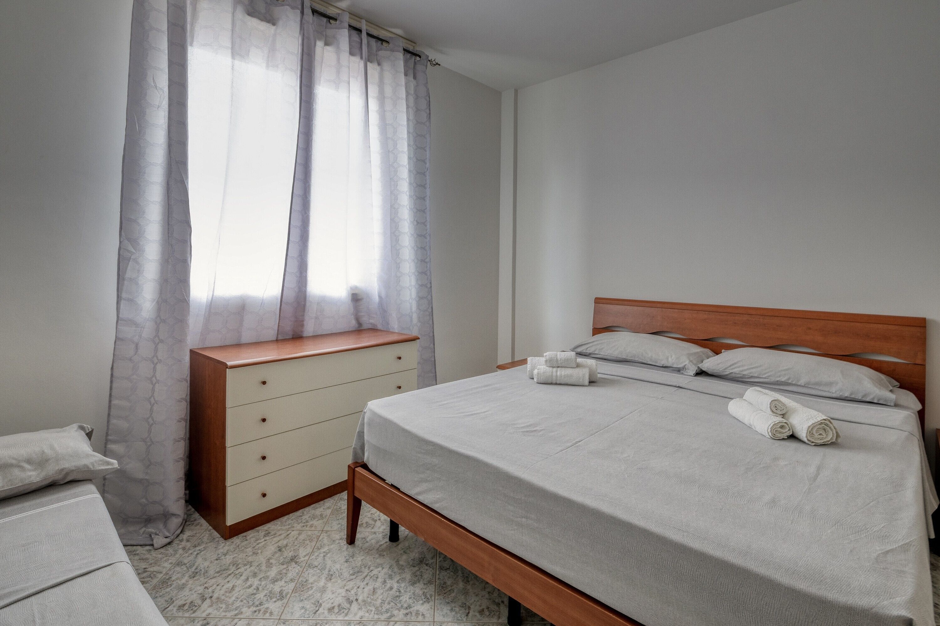 Una camera da letto con un letto, una cassettiera in legno, una finestra con tende e un pavimento piastrellato.