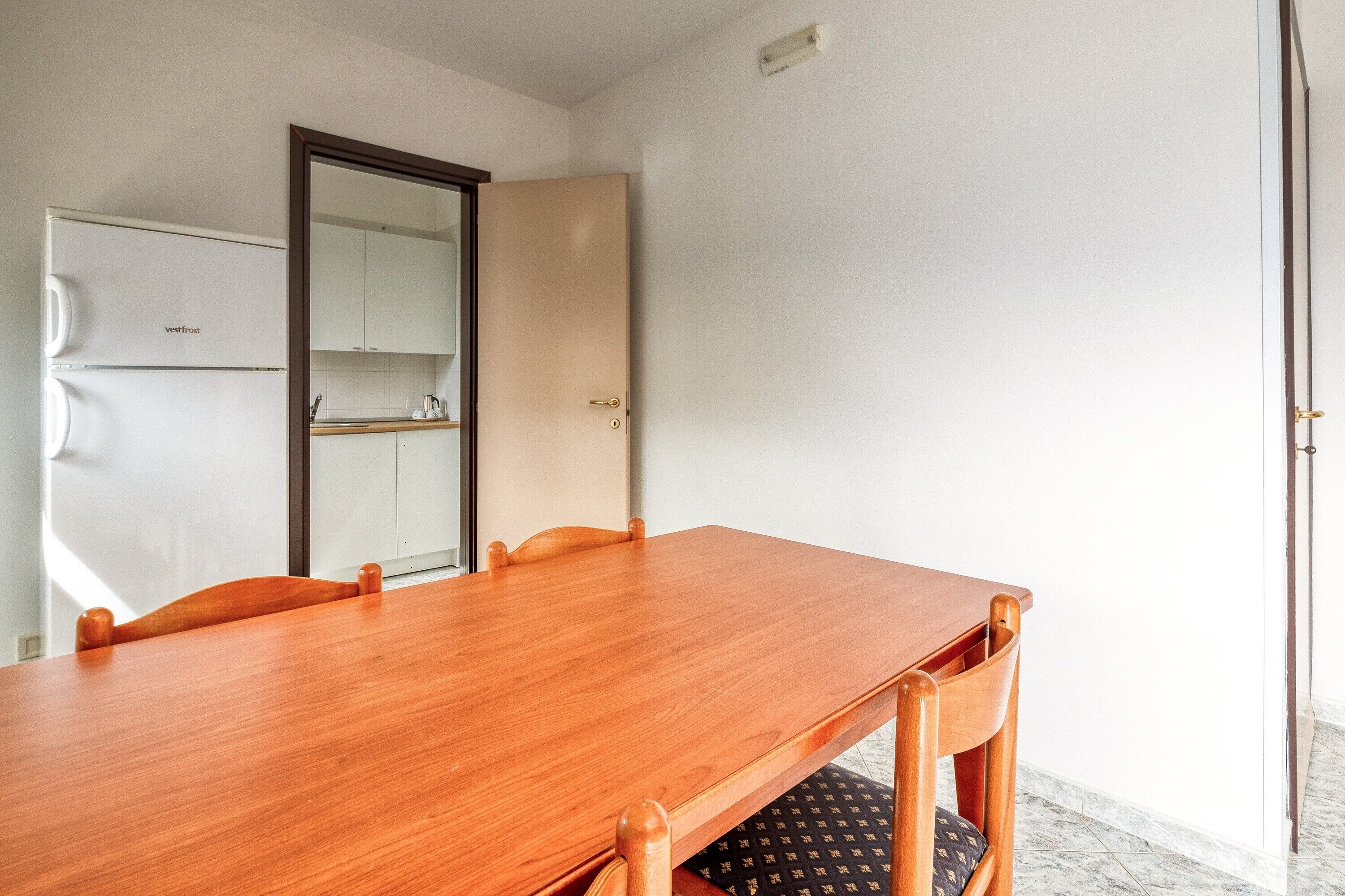 A room with a wooden dining table, chairs, a white refrigerator, and a door.