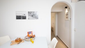 En matplats med ett vitt bord, en korg med bröd, en påse chips och två glas juice. På väggen hänger två svartvita fotografier.