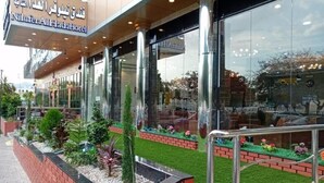 A hotel entrance with a sign in Arabic and English, a glass facade, and a landscaped garden area. - Nilufer AlHada Hotel (Taif)