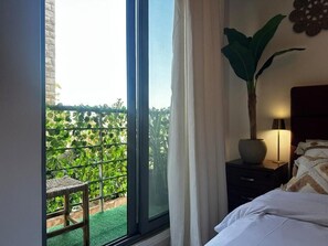 A hotel room with a large window, a bed, a nightstand, a lamp, and a potted plant. - Boho Studio (Amman)