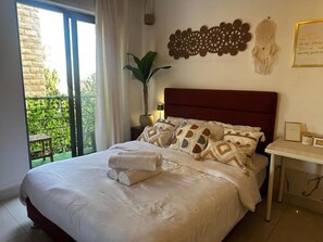 A bedroom with a large bed, a nightstand, a wall decoration, and a view of greenery outside. - Boho Studio (Amman)