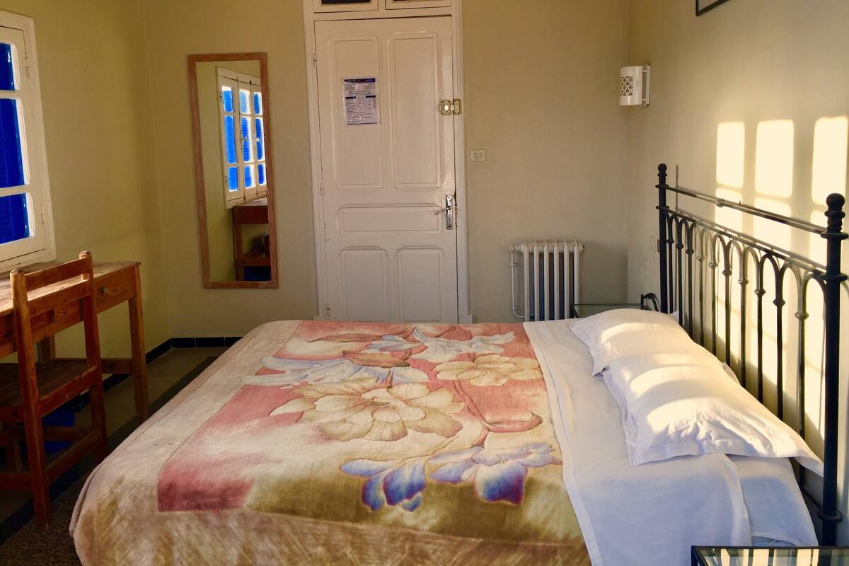 A bedroom with a bed, a wooden chair, a white door, and a window with blue shutters.