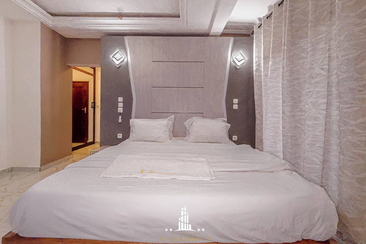 Une chambre spacieuse avec un grand lit, une tête de lit ornée de motifs géométriques et une porte visible donnant sur une autre pièce.