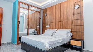 A modern bedroom with a large bed, wooden panel walls, a mirror, and a nightstand. - FT Residence (Douala)