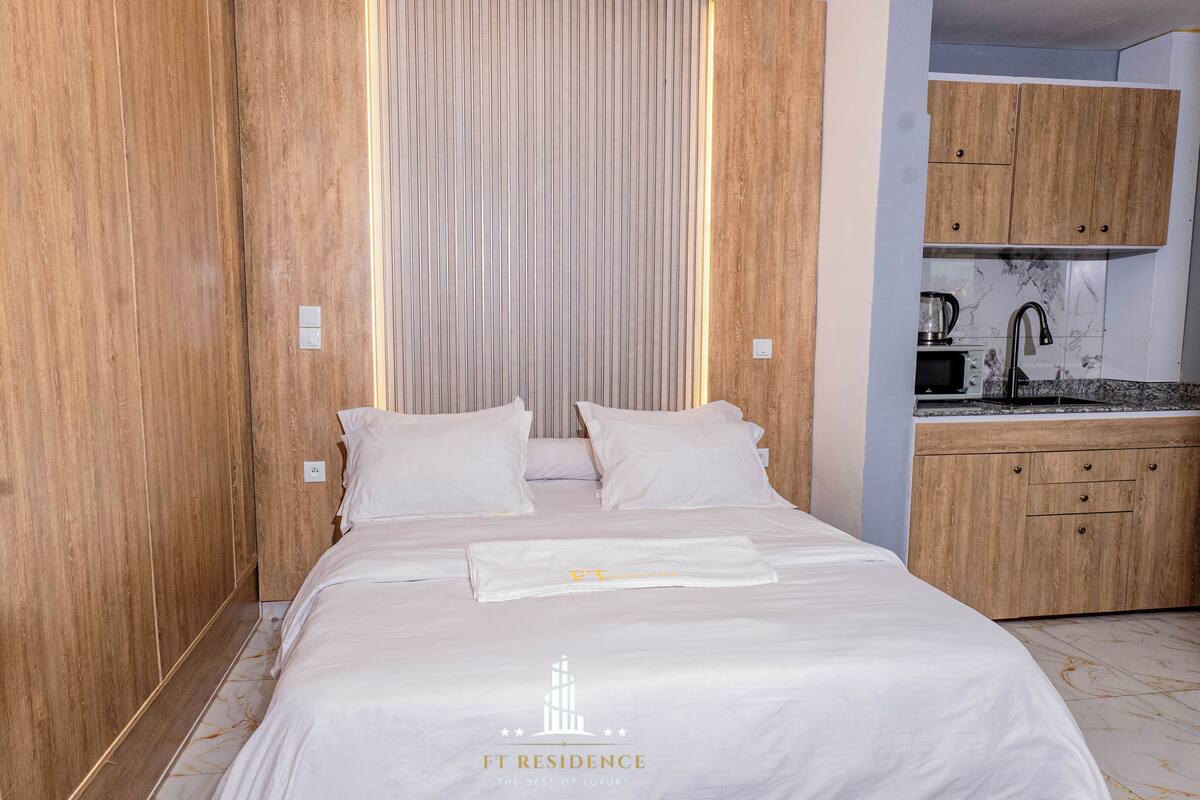 Une chambre à coucher comprenant un lit, des oreillers, une table de chevet, une armoire en bois et une kitchenette équipée d’un évier et d’un four à micro-ondes.