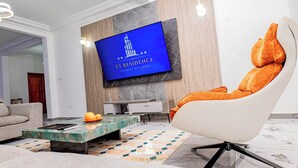 A modern living room with a large flat-screen TV mounted on the wall, a comfortable orange chair, a marble coffee table, and a patterned rug. - FT Residence (Douala)