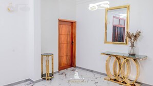 A hallway with a wooden door, a round gold table with a vase, and a mirror on the wall. - FT Residence (Douala)