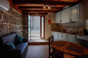 A cozy interior with a stone wall, a wooden ceiling, a sofa with cushions, a dining table, and a kitchen area with cabinets and a sink. - O Palheiro - Casas Vale do Laboreiro (Melgaço)