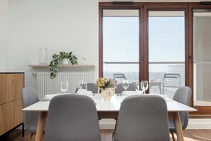 Ein Essbereich mit einem Tisch für zwei Personen, einer Topfpflanze auf einem Regal und Blick durch ein großes Fenster.