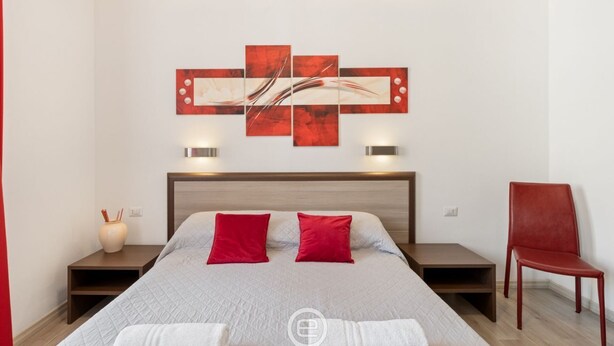 Una habitación de hotel con una cama, una silla roja, una mesita de noche, una obra de arte en la pared y un jarrón.