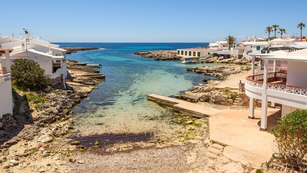 A coastal town with clear blue waters, rocky shoreline, and white buildings with balconies.