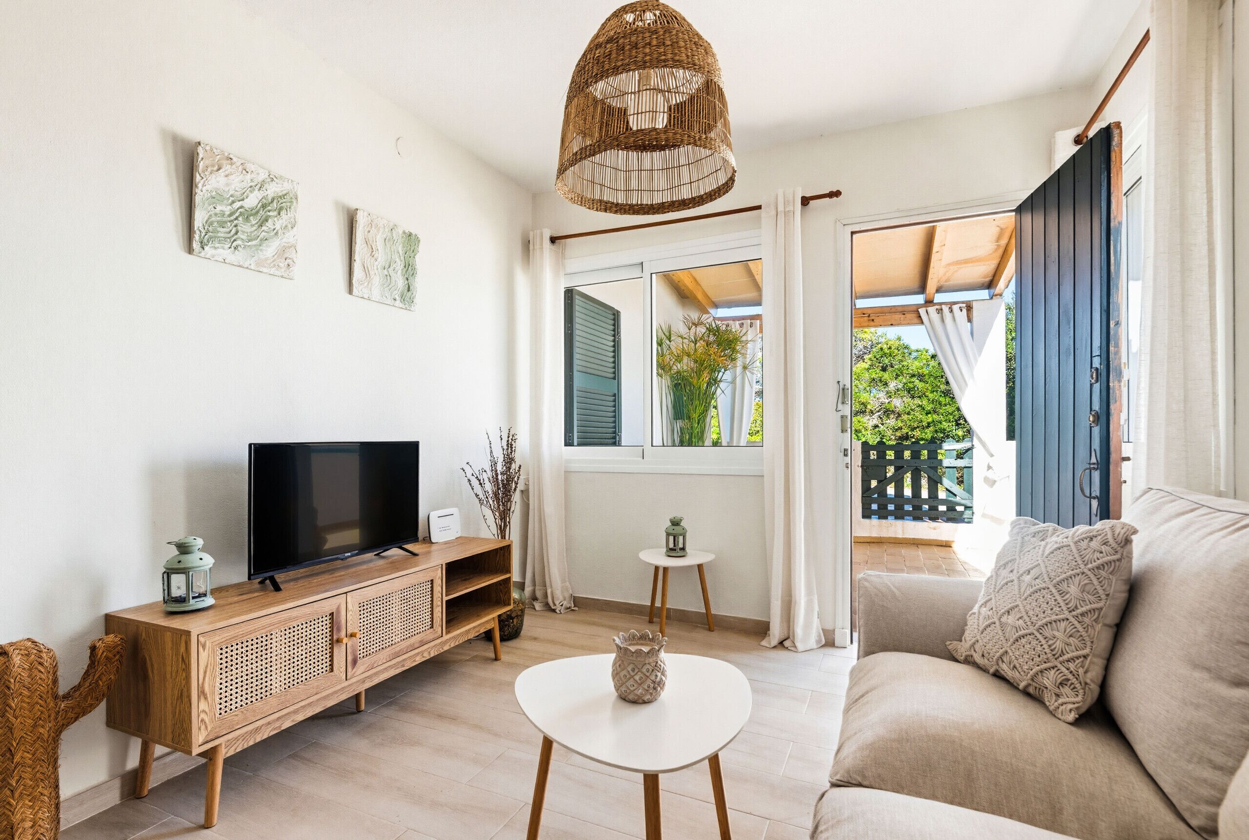 A living room with a TV, a white coffee table, a sofa, a wicker lamp, and a view of the outside.