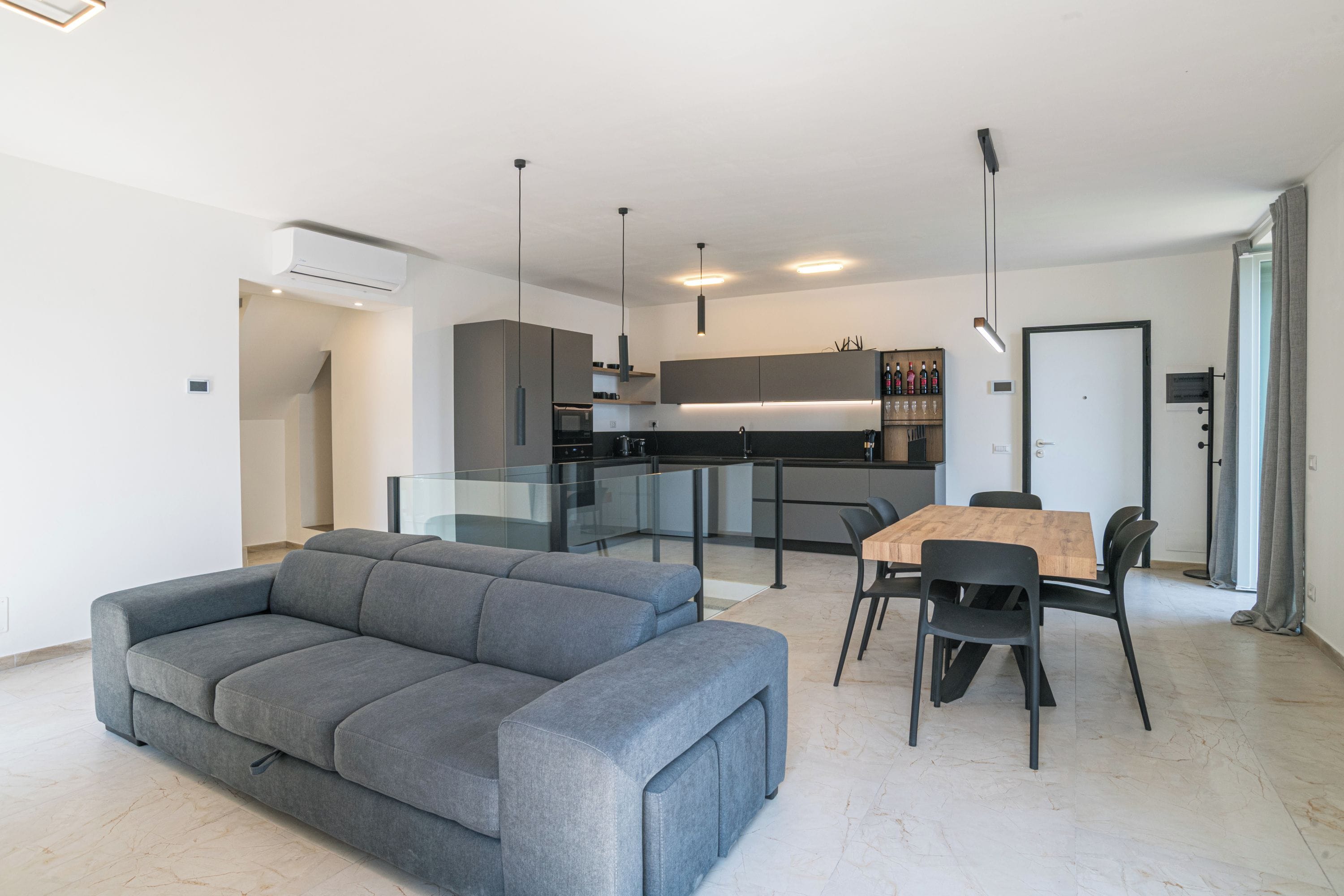 A modern living room with a grey sofa, a dining table with chairs, and a kitchen area in the background.