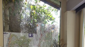 Ein Zimmer mit einer großen Karte an der Wand, einem kleinen Tisch mit Reisebroschüren und einem Fenster mit Blick auf Bäume und Gebäude.