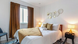 En pænt redt seng med hvid dyne og brun plaid, et natbord med lampe, samt en dekorativ vægplade.