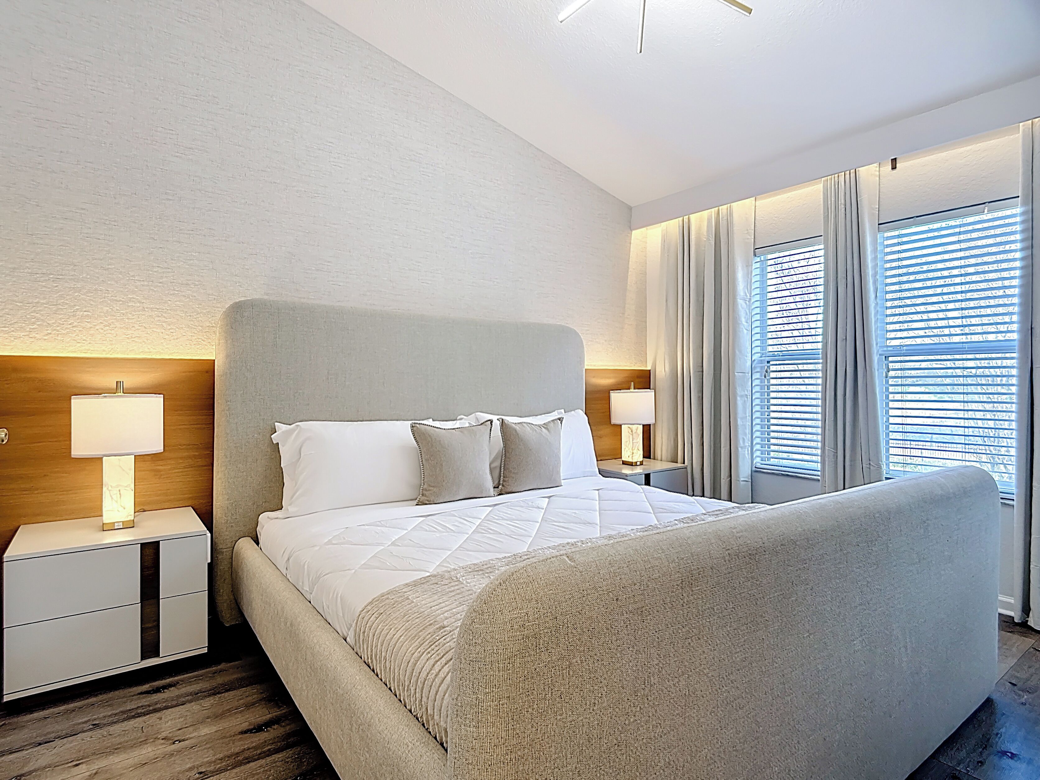 Une chambre d’hôtel moderne avec un grand lit, deux tables de chevet avec des lampes, une tête de lit en bois et une grande fenêtre avec des stores.