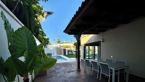 Une terrasse avec une piscine, un espace repas couvert et de grandes plantes vertes.
