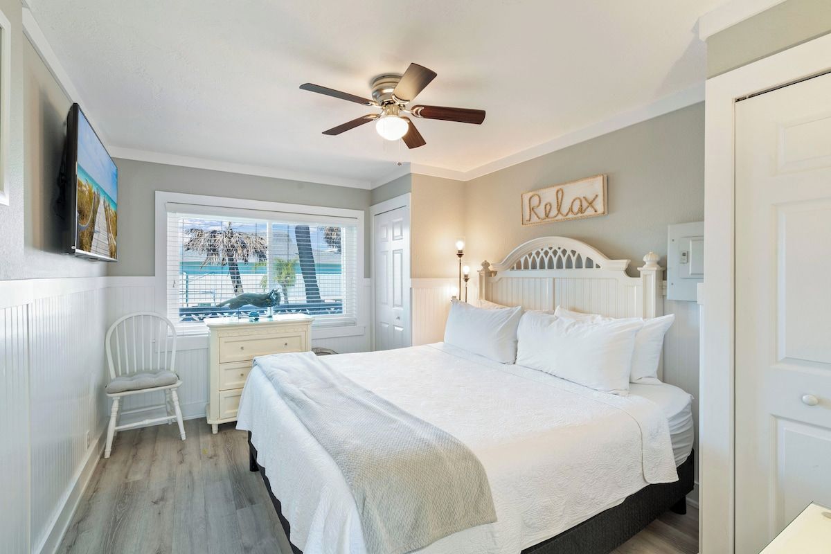 A bedroom with a bed, a desk, a chair, a ceiling fan, and a window with a view.