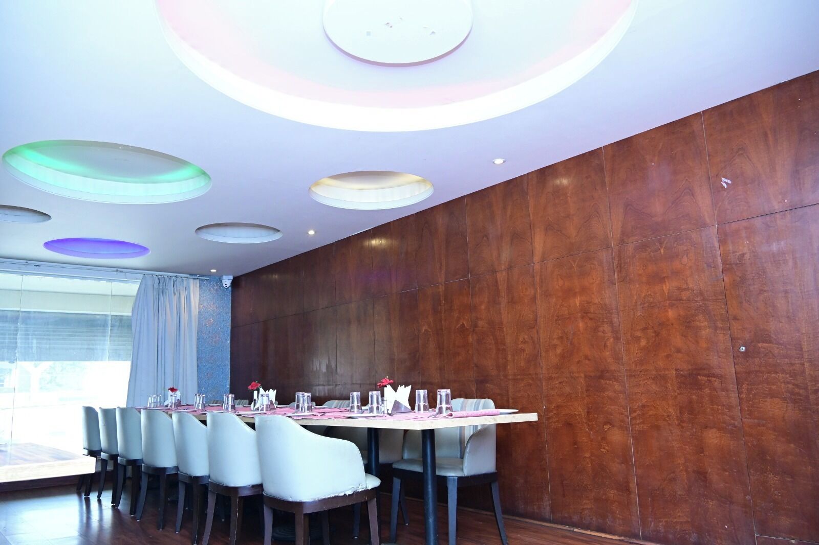 Un espace repas avec une longue table dressée pour six personnes, entourée de chaises, avec un mur en bois et un plafond orné de luminaires circulaires.