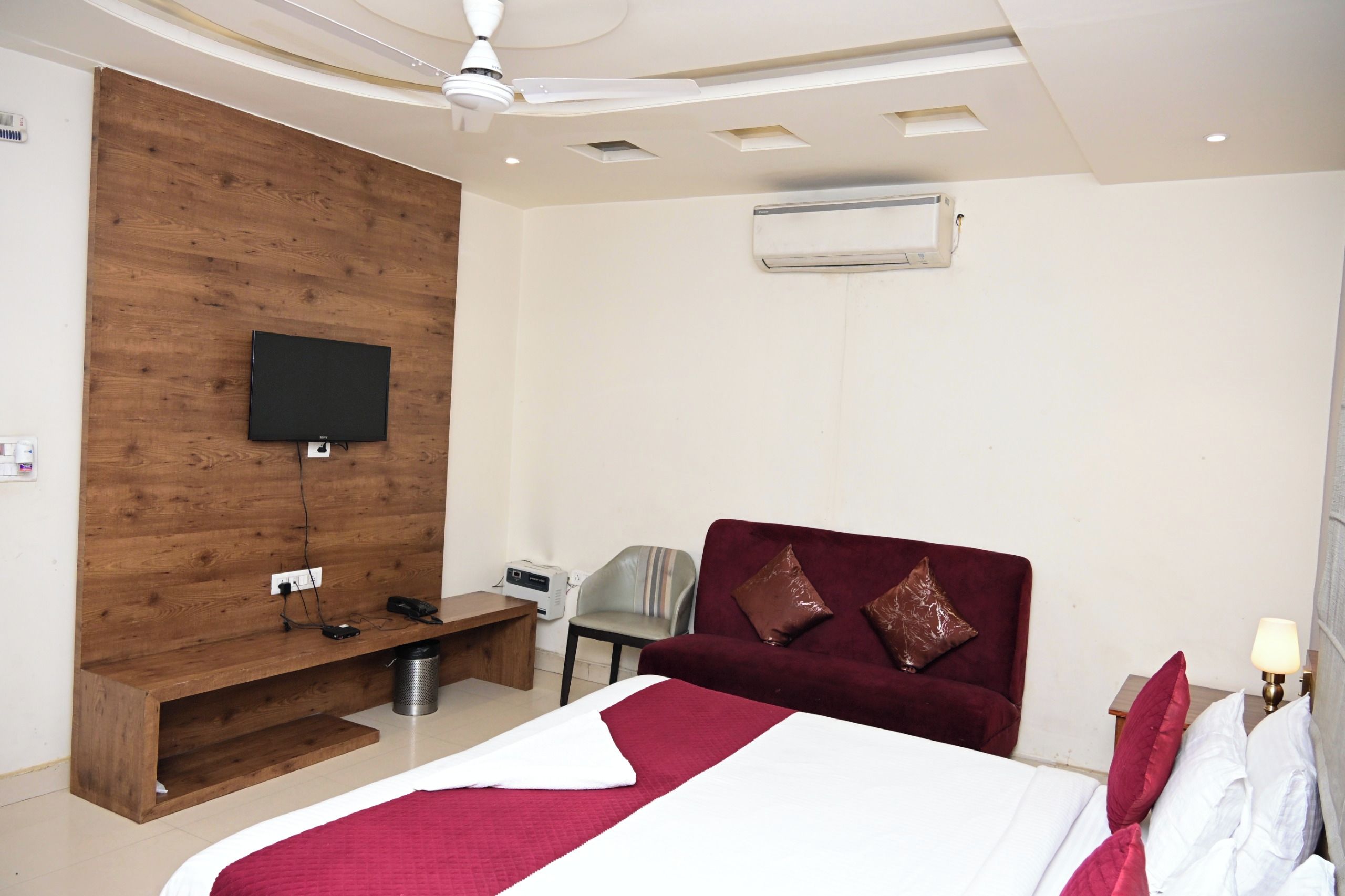 A hotel room with a bed, a TV mounted on a wooden wall, a ceiling fan, and a window with an air conditioner.