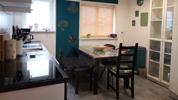 Een moderne keuken met een centraal eiland, een eettafel met stoelen, een raam met zonwering, en een kast met glazen deuren.