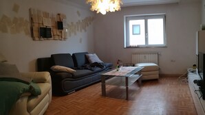 Een woonkamer met een bank, een salontafel, een televisie en een raam met zonwering.