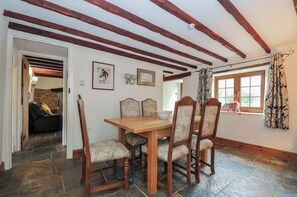 A dining area with a wooden table and chairs, stone flooring, exposed wooden beams, and a window with curtains. - Little Blagdon (Holsworthy)